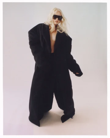 Model with long blonde hair wearing oversized black coat and sunglasses against plain background