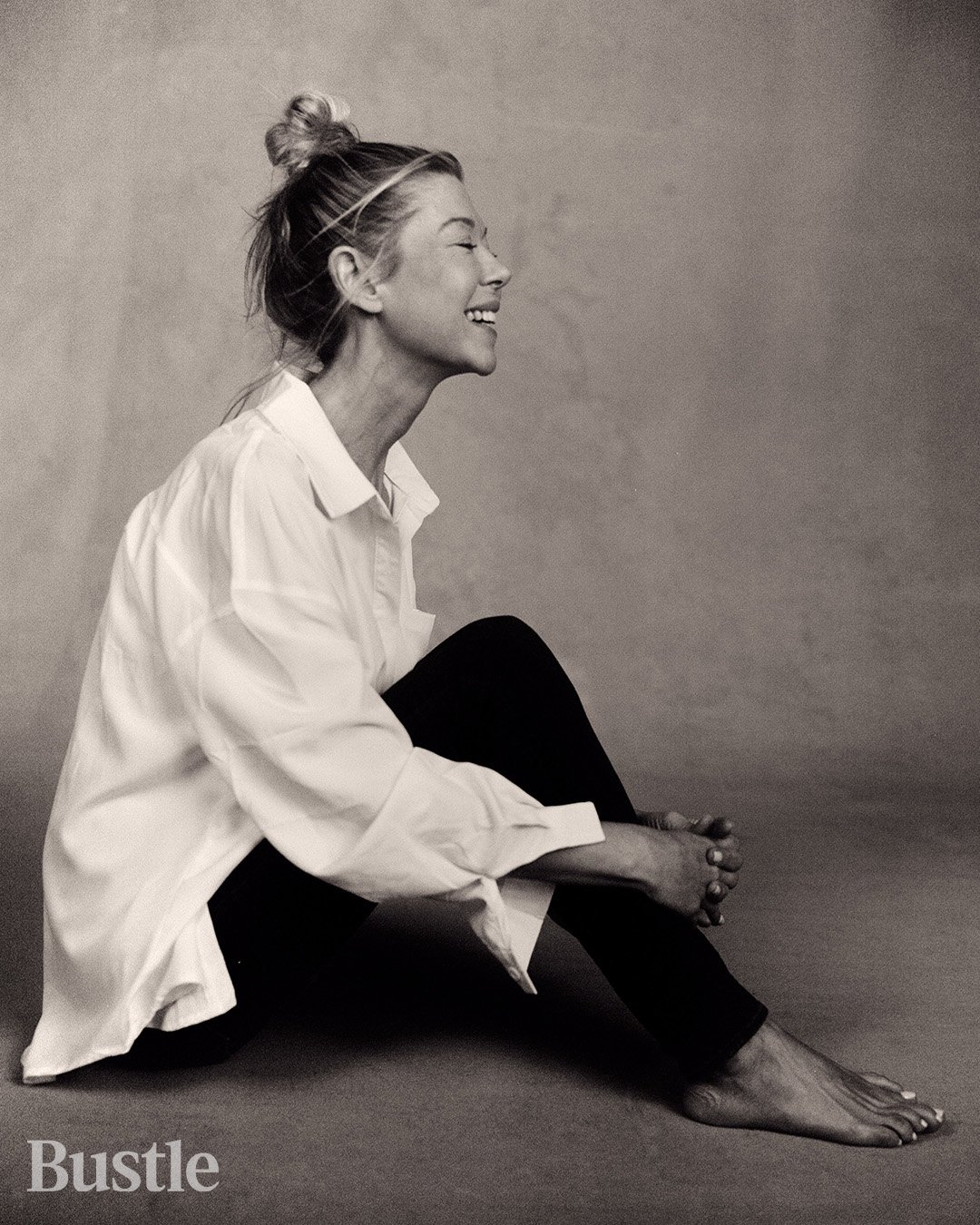 Black and white portrait of a smiling adult woman with hair in a bun, wearing a white shirt and black pants, sitting on the floor.