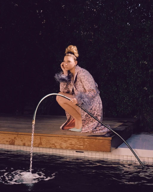 Young woman in floral robe sitting at edge of pool with metal water feature. She is resting her head in her hand in thought.