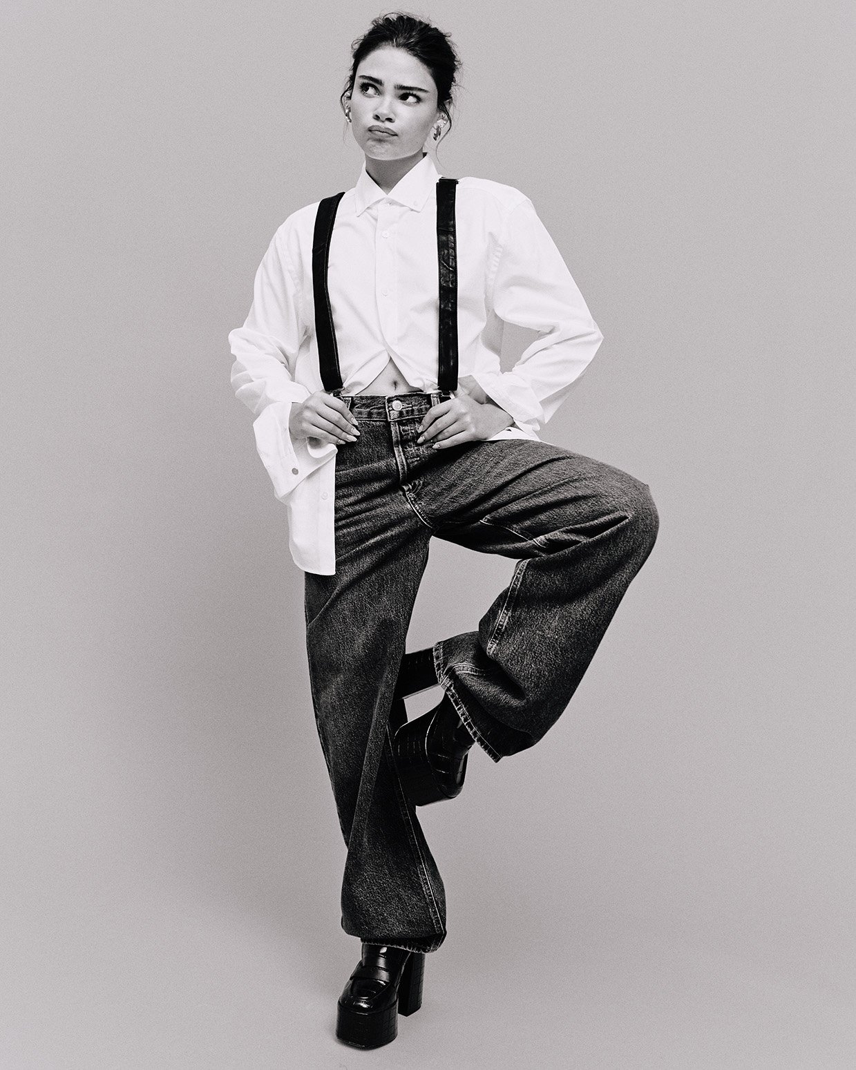 Monochrome full shot of a young adult female in a white shirt, suspenders, and jeans standing on one leg in a neutral studio.