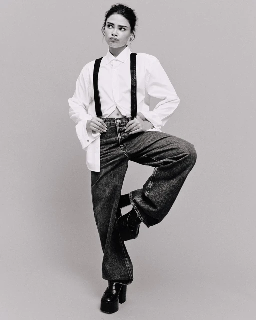 Monochrome full shot of a young adult female in a white shirt, suspenders, and jeans standing on one leg in a neutral studio.