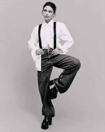 Monochrome full shot of a young adult female in a white shirt, suspenders, and jeans standing on one leg in a neutral studio.