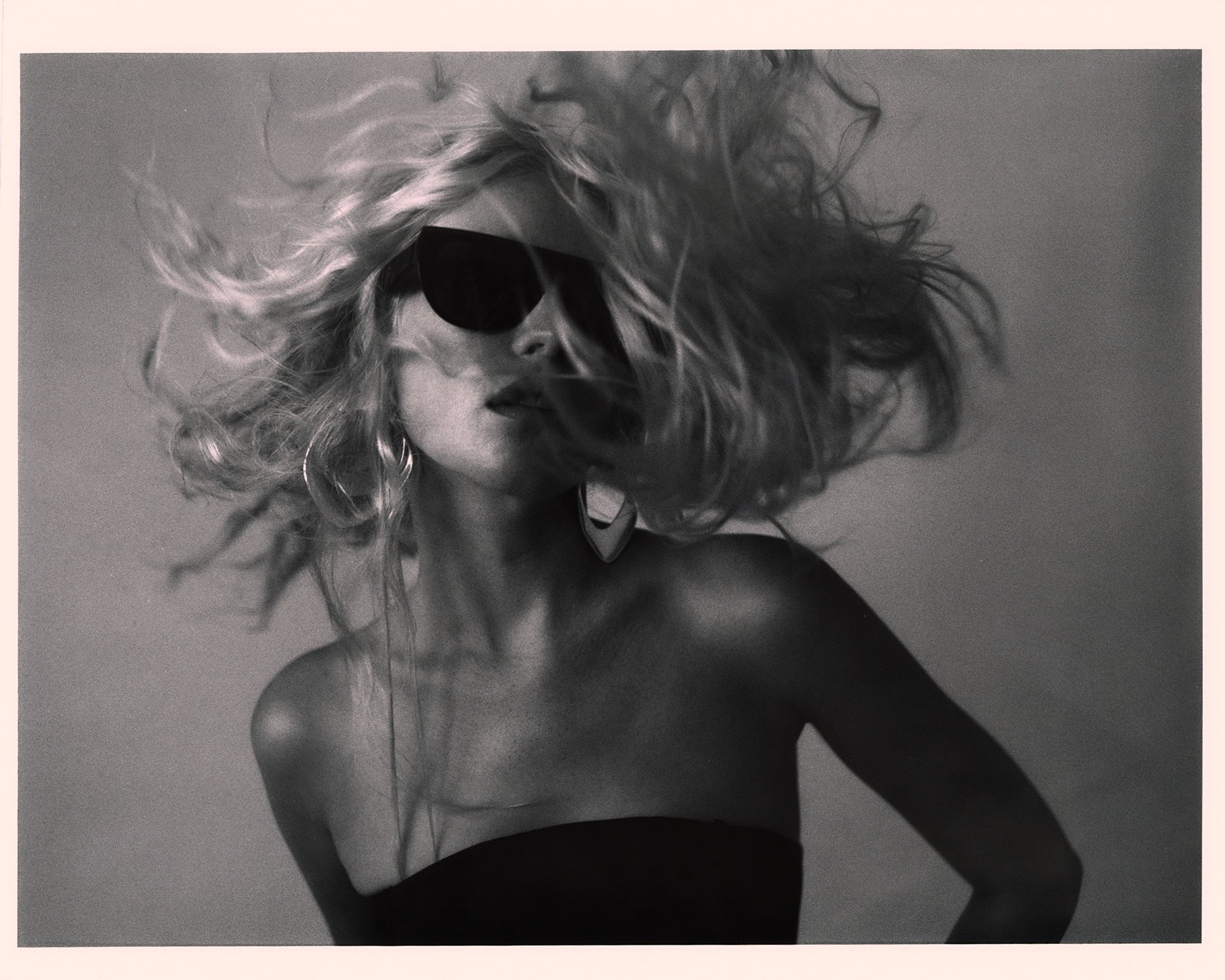Monochrome portrait of a blonde woman with sunglasses. Studio lighting enhances the dramatic wind-swept hair and earring details.