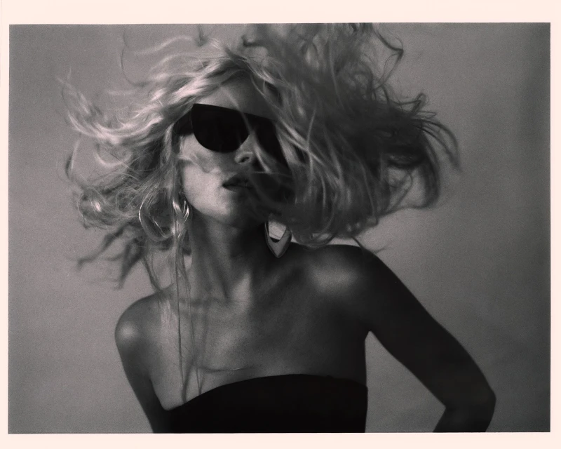 Monochrome portrait of a blonde woman with sunglasses. Studio lighting enhances the dramatic wind-swept hair and earring details.