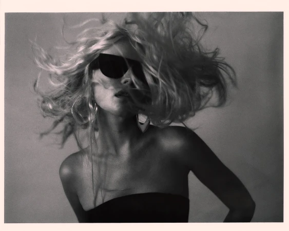 Monochrome portrait of a blonde woman with sunglasses. Studio lighting enhances the dramatic wind-swept hair and earring details.