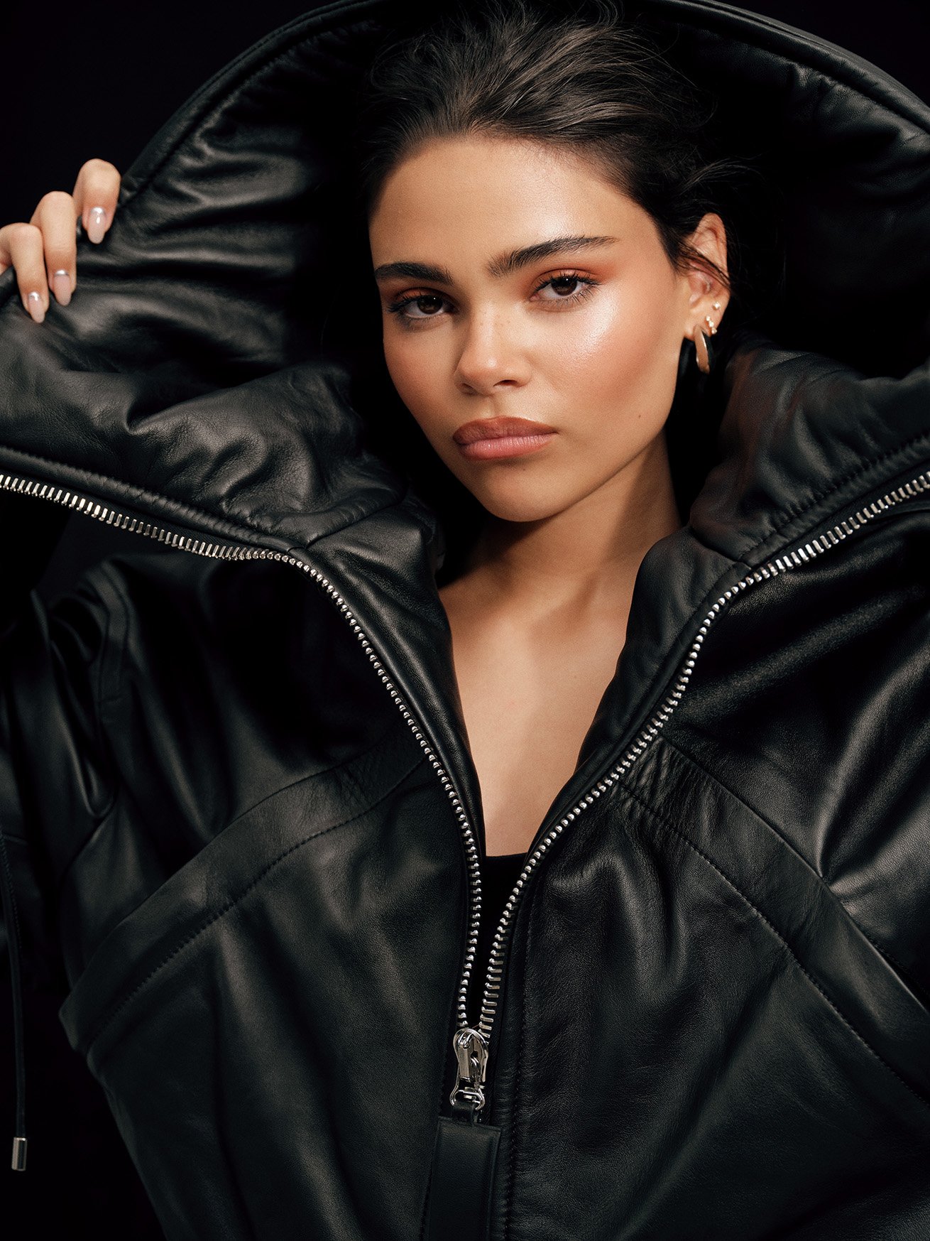 Close-up of a woman with neutral expression wearing a black hooded leather jacket, photographed in a studio setting with a dark background.
