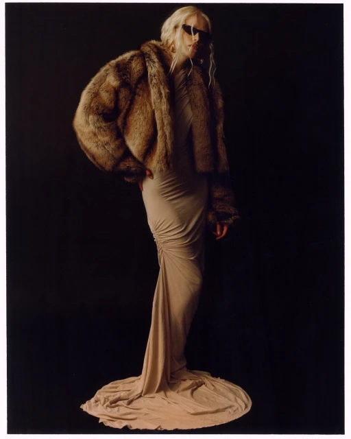 Fashionable woman in beige gown and fur coat posing against dark backdrop in studio lighting, exuding elegance and modern style.