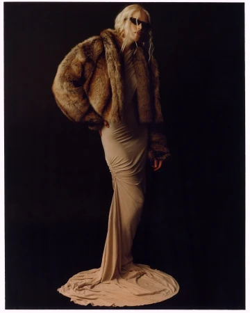 Fashionable woman in beige gown and fur coat posing against dark backdrop in studio lighting, exuding elegance and modern style.