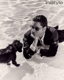 Man wearing sunglasses and a dark suit lying in a shallow swimming pool looking at a small black dog by Alex G. Harper