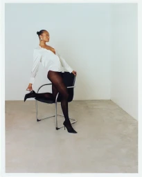 Woman in white shirt and black tights poses leaning on a modern office chair in a minimalist white room