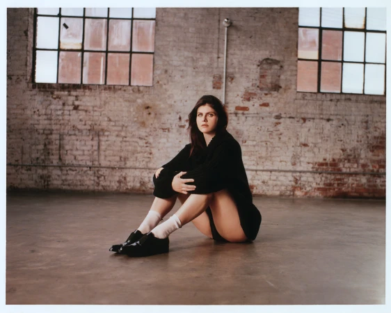 Alex Daddario seated on a concrete floor wearing a black sweater, white socks, and black shoes against a brick wall.