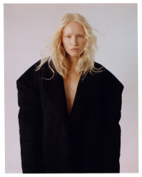 Portrait of a blonde woman wearing an oversized black coat against a plain light background