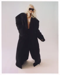 Model with long blonde hair wearing oversized black coat and sunglasses against plain background
