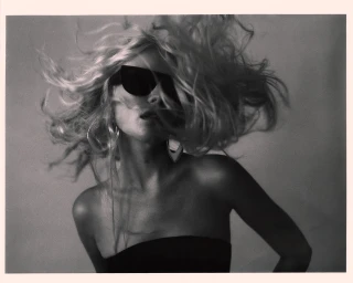 Monochrome portrait of a woman with flying blonde hair wearing dark sunglasses and hoop earrings against a neutral background.