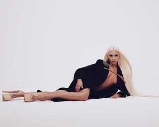 Model with long blonde hair wearing a black coat and platform heels lying on a white seamless background