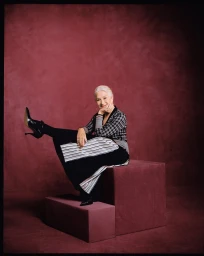 Helen Mirren seated on a burgundy block wearing a plaid cardigan and black skirt with her leg raised playfully.