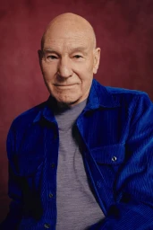 Portrait of Patrick Stewart wearing a royal blue corduroy shirt and grey t-shirt against a textured dark red background.