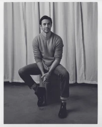 Portrait of a man in a crewneck sweater and cuffed jeans seated against a draped curtain background by Alex G. Harper