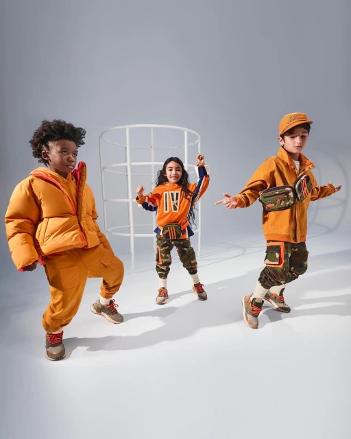 Three children pose in orange and camouflage outfits against a solid grey backdrop. The kids are wearing matching streetwear.