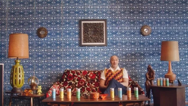 A bald man in an orange vest and blue pants sits on a floral couch with colorful drink cans on the table in front of patterned wall.