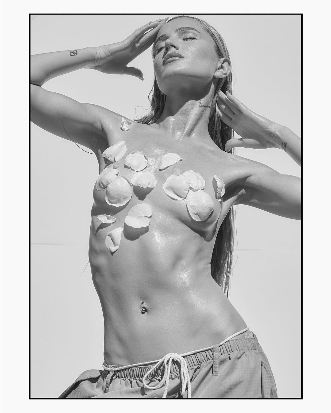 Black-and-white portrait of a woman with flower petals on her torso, hands above her head, eyes closed, and a light background.