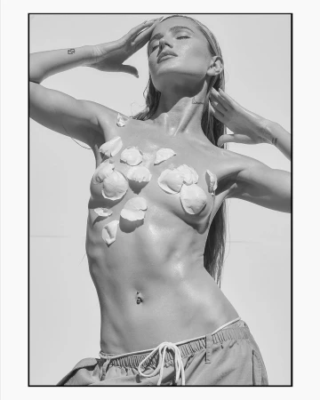 Black-and-white portrait of a woman with flower petals on her torso, hands above her head, eyes closed, and a light background.