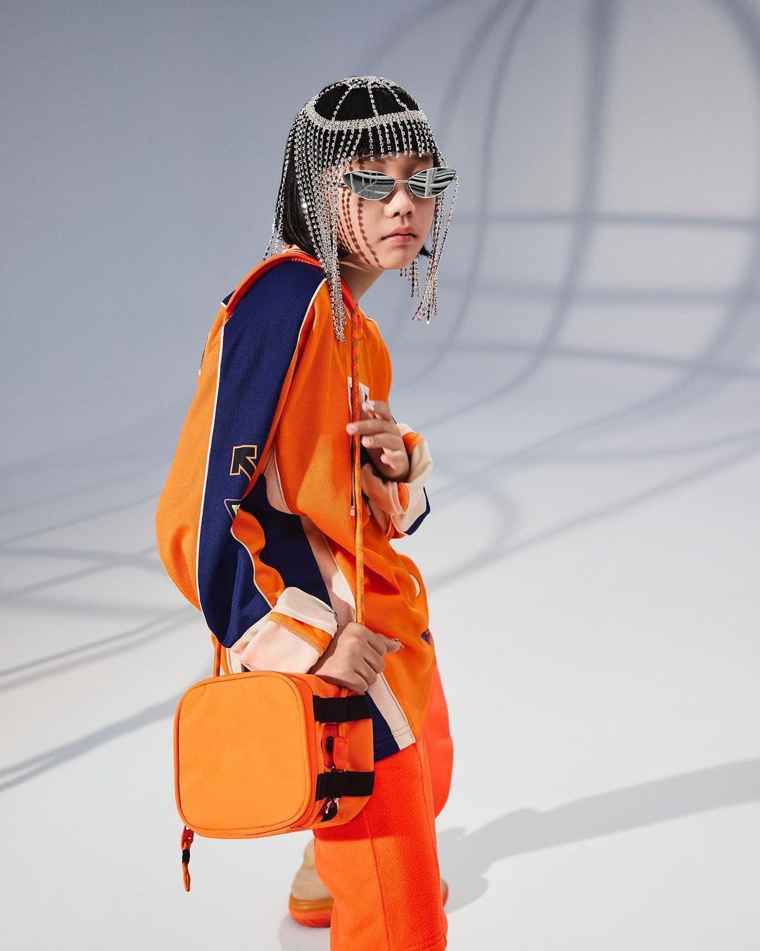 Child model in an orange outfit, wearing a beaded headpiece and sunglasses, poses against a light gray background holding a purse.