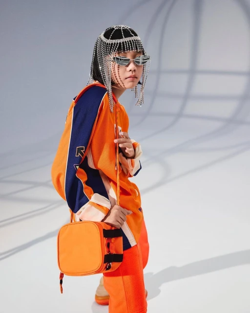 Child model in an orange outfit, wearing a beaded headpiece and sunglasses, poses against a light gray background holding a purse.