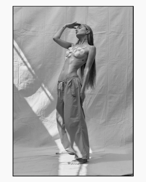 Black and white portrait of a slender woman with long hair, wearing floral accents and loose pants, posing in front of textured white backdrop.