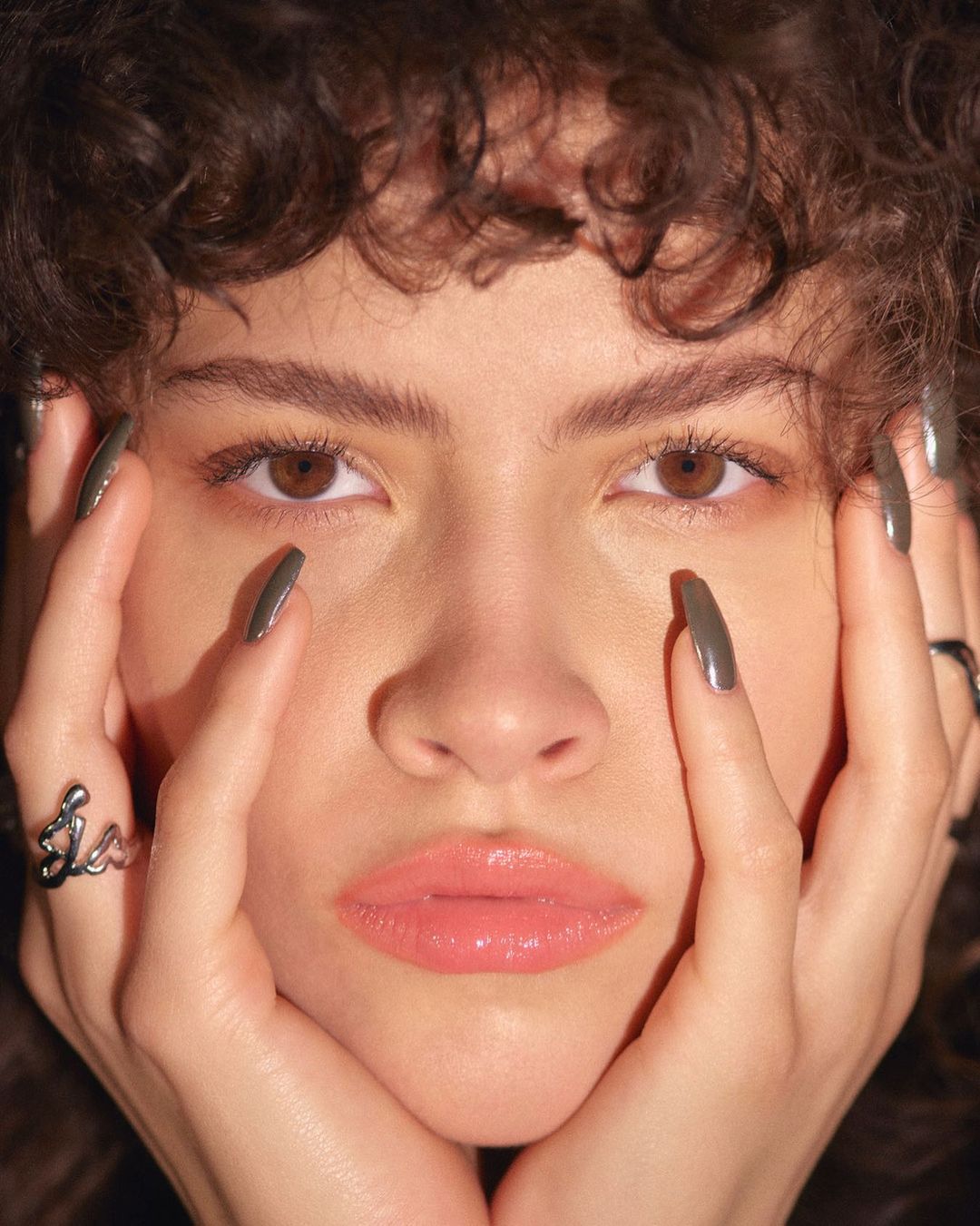 Portrait of a young adult woman with curly brown hair, brown eyes, and gray nail polish, her face framed by hands with statement rings.