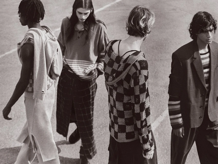 Four fashion models stand outdoors in streetwear-inspired outfits. The image is black and white with a shallow depth of field.