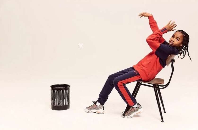 Young person sits backward on chair, tossing crumpled paper toward black wire trash can on white backdrop in a fashion shot.