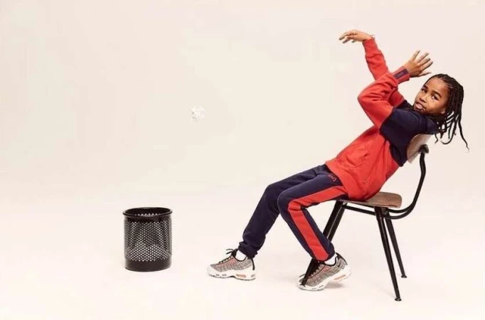 Young person sits backward on chair, tossing crumpled paper toward black wire trash can on white backdrop in a fashion shot.