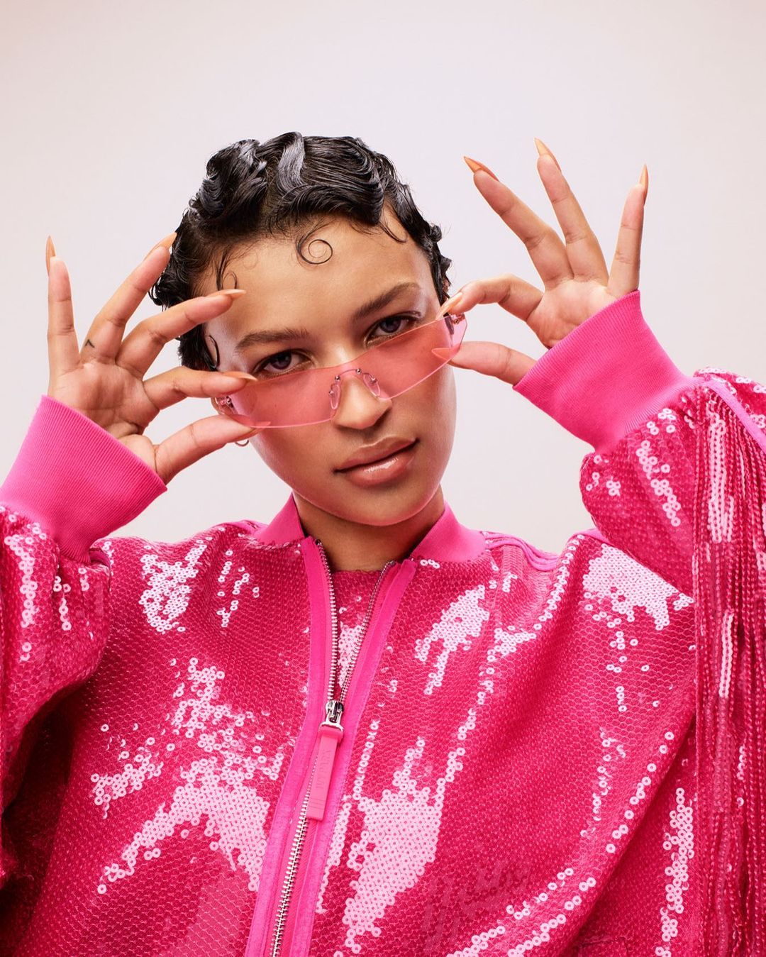 Woman with dark, wavy hair and pink sunglasses in pink, sequined bomber jacket smiles in an eye-level studio shot.