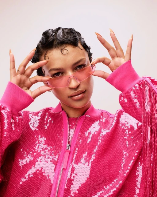 Woman with dark, wavy hair and pink sunglasses in pink, sequined bomber jacket smiles in an eye-level studio shot.