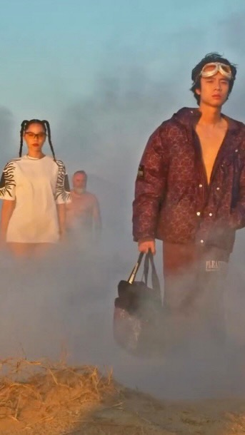 Three people walk through a hazy outdoor setting, with a man in a maroon jacket and goggles carrying a duffel bag in the foreground.