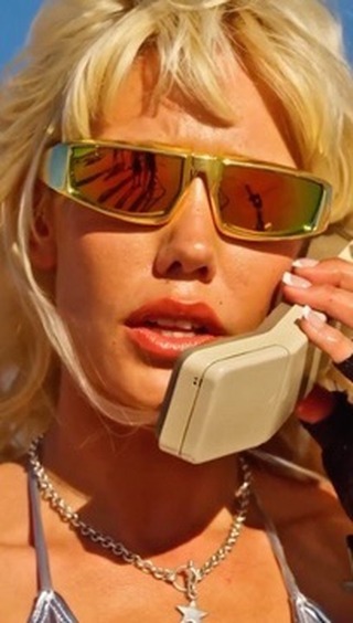Close-up of an adult female with blonde hair talking on a landline phone, wearing sunglasses and a silver-colored top, outdoors.