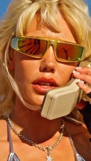 Close-up of an adult female with blonde hair talking on a landline phone, wearing sunglasses and a silver-colored top, outdoors.