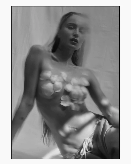 Black and white studio shot of a woman with long hair and flower petals on her chest, against a draped fabric backdrop with soft lighting.