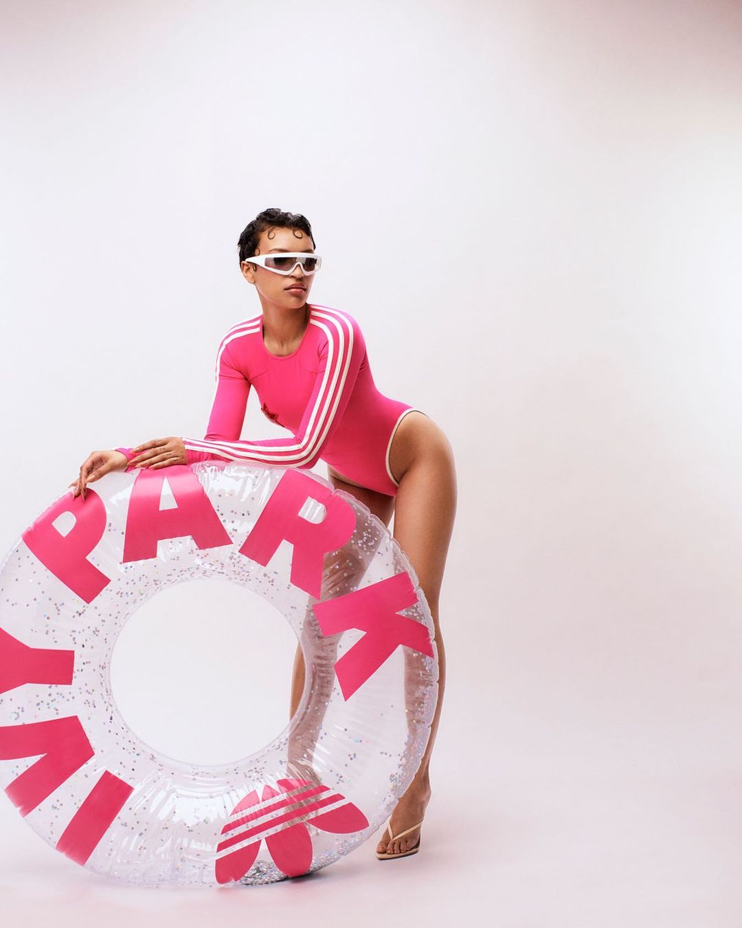 Woman in pink swimsuit posing with Ivy Park inflatable float against white background. Fashion shoot features Adidas branding and sunglasses.