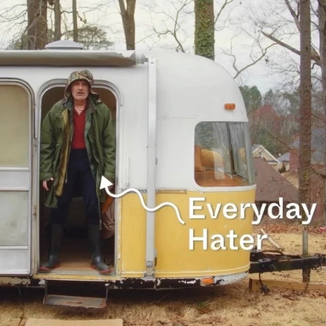 A man in an outdoor outfit standing in the doorway of a vintage travel trailer, with trees and a house in the background.
