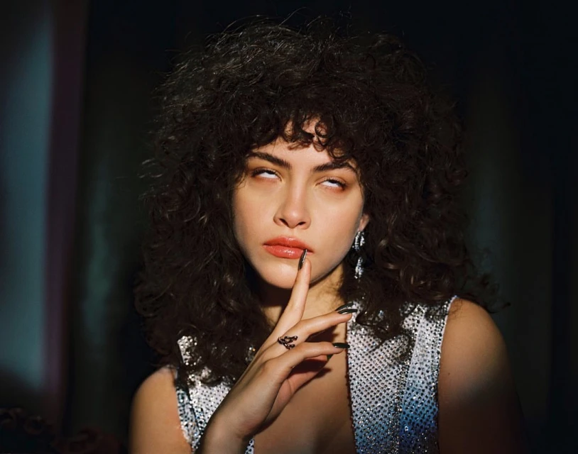 Close-up studio portrait of a young adult woman with curly hair, shiny sleeveless top, finger to lips, and a mischievous facial expression.