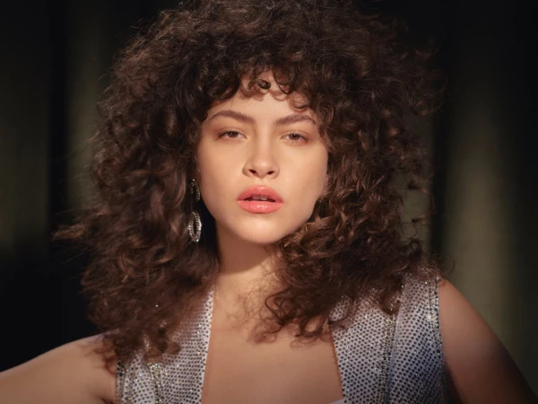 Portrait of a woman with voluminous curly dark hair wearing a sparkling embellished vest and hoop earrings.