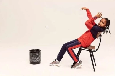 Young boy in red and blue tracksuit leaning back on a metal chair aiming a paper ball at a trash can