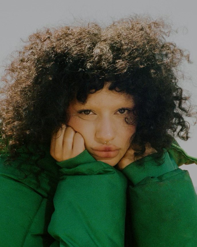 Portrait of a woman with dark curly hair and a septum ring wearing a green jacket, resting her chin on her hands.