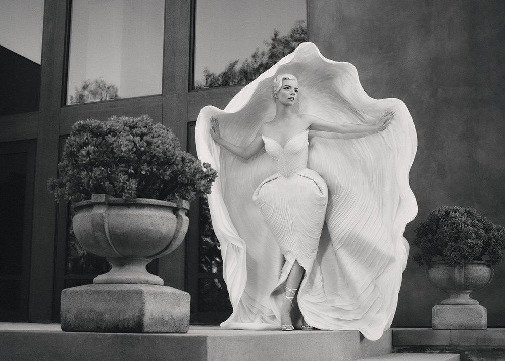 Woman in white gown poses on stairs between matching planters and a modern building. Black and white fashion, architectural, portrait photograph.