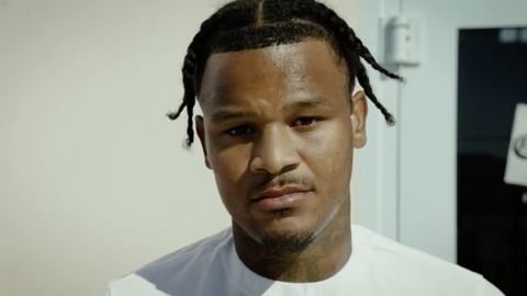 Portrait of a young adult male with braided hair, wearing a white shirt, with a neutral expression, captured in an indoor setting.
