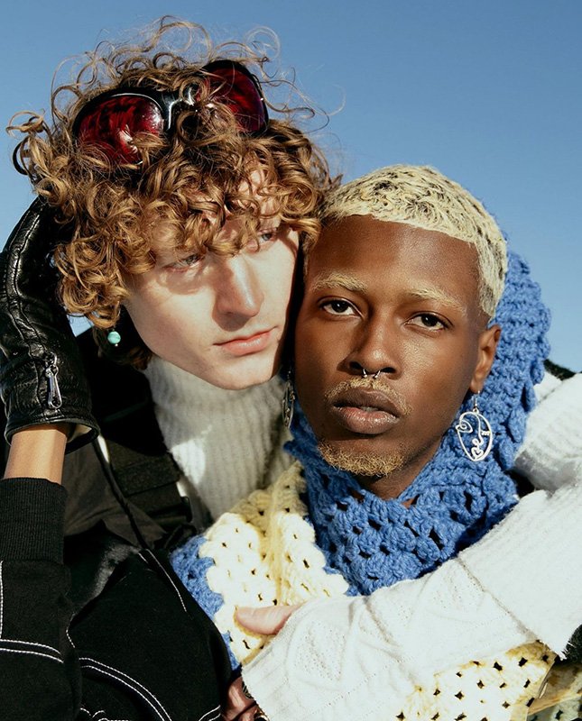 Two fashionable young adults in a close-up shot. One person has curly hair and sunglasses, while the other has bleached blonde hair.