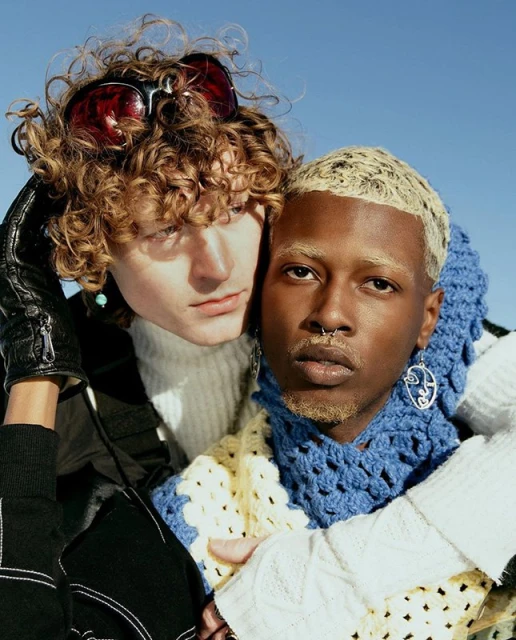 Two fashionable young adults in a close-up shot. One person has curly hair and sunglasses, while the other has bleached blonde hair.