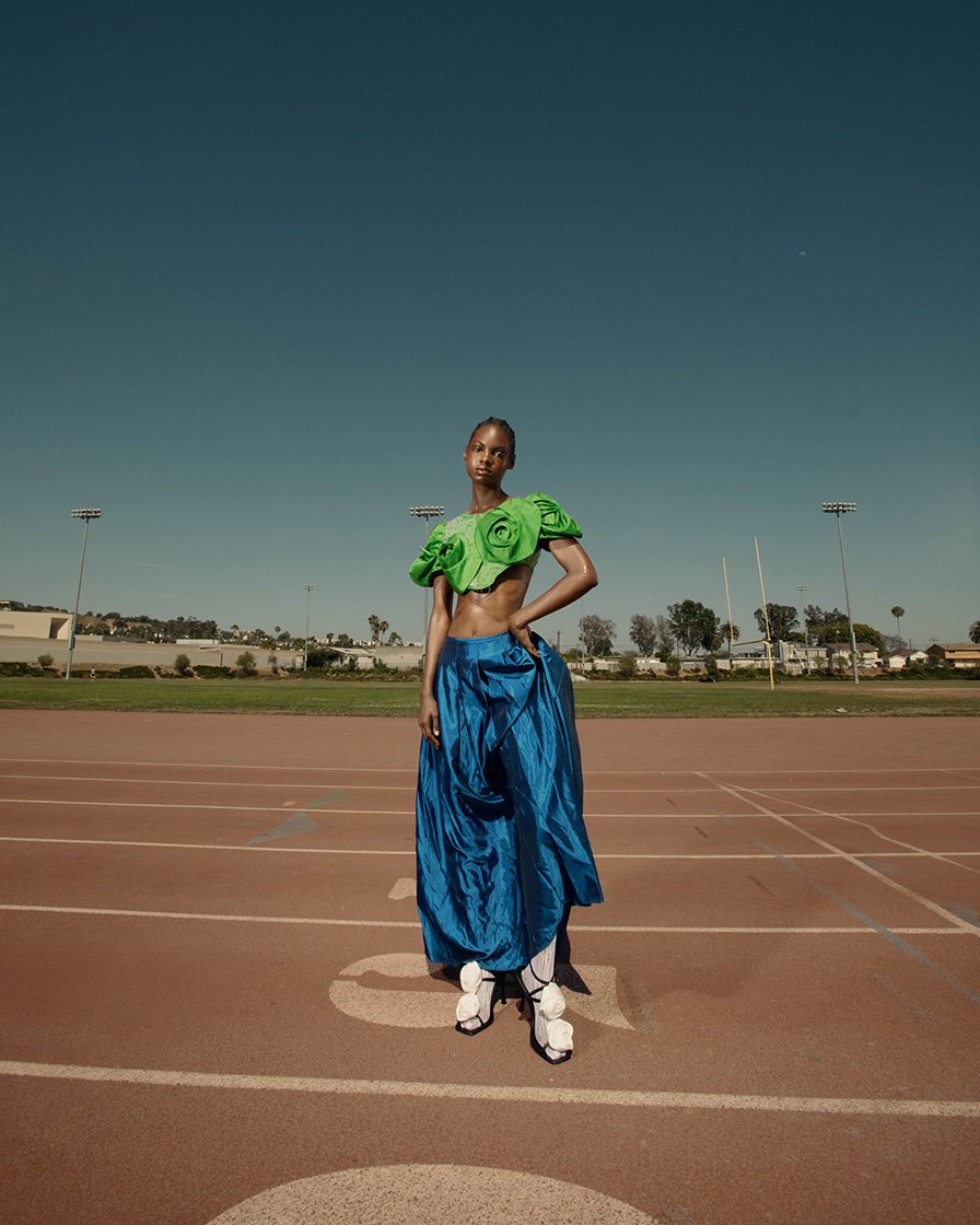 Fashion portrait of a person on a running track with a bright green top and shiny blue skirt on a sunny day with blue sky.